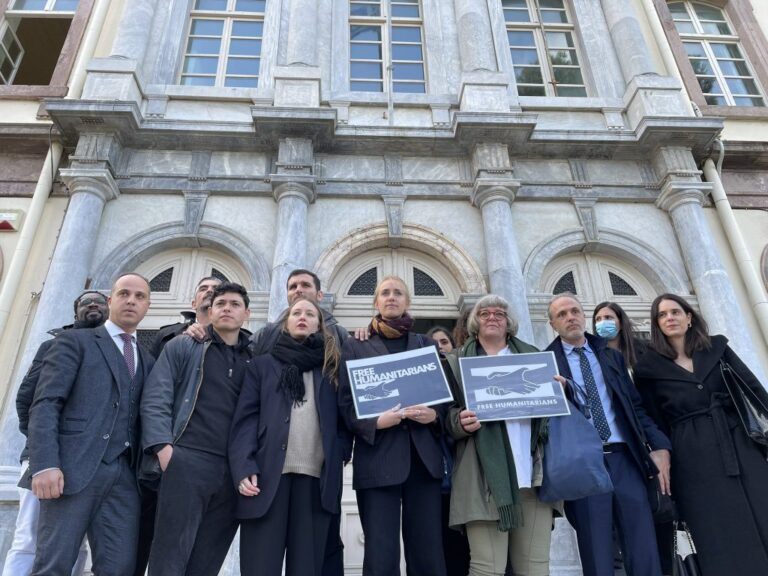 Photo: This morning, Seán Binder stands alongside other defendants and their legal defense team, holding up banners demanding the freedom of relief workers from the steps of the courthouse where the trial is being held on the Greek island of Lesbos. © Posted by Begüm Başdaş on Twitter.