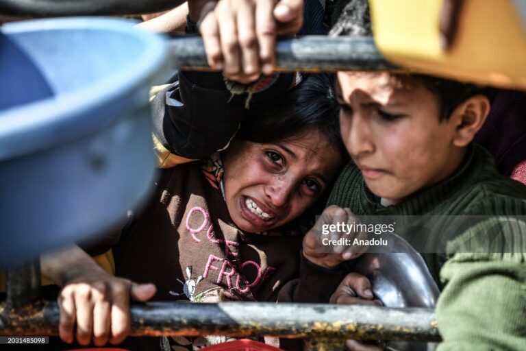 رفح، غزة - 25 فبراير/شباط: طفل عالق بينما يحمل الفلسطينيون أوعيتهم الفارغة لتعبئتها بالمواد الغذائية التي توزعها المنظمات الخيرية خلف القضبان لعدم قدرتهم على الحصول على المواد الغذائية الأساسية بسبب الحظر الذي تفرضه القوات الإسرائيلية في رفح، غزة، في 25 فبراير/شباط 2024. (تصوير عابد زقوت/الأناضول عبر غيتي)